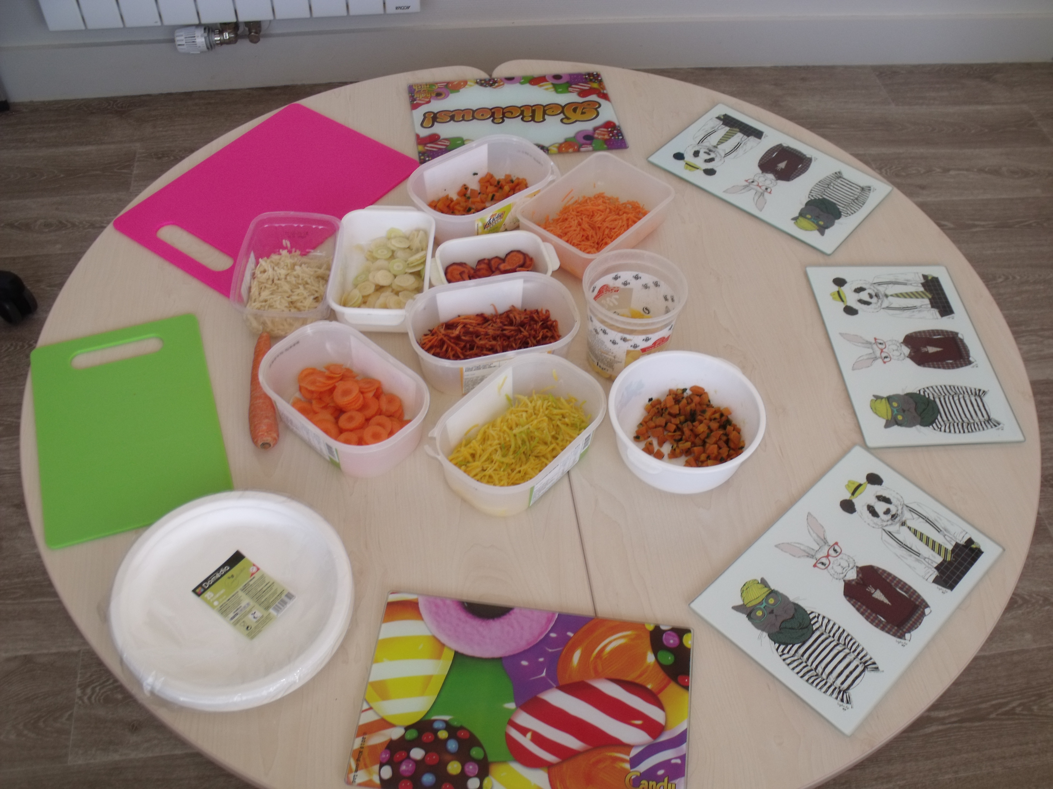 Sur une table ronde à hauteur d’enfants, des assiettes et des planches en verre décorées, sont disposées. Au centre se trouvent des ramequins avec des carottes sous toutes les formes : en julienne, râpées, en petits morceaux, en entière, cuites ou crues et de trois couleurs.