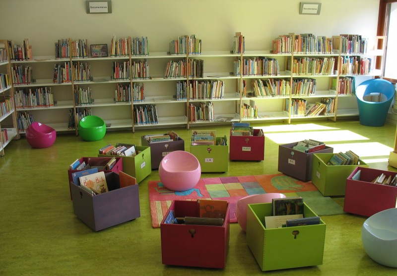 Dans la salle qui accueille les plus petits, des tabourets de formes arrondis et de toutes les couleurs sont installés autour d’un tapis coloré. Des caisses de livres sur roulettes sont disposés en rond autour du même tapis.