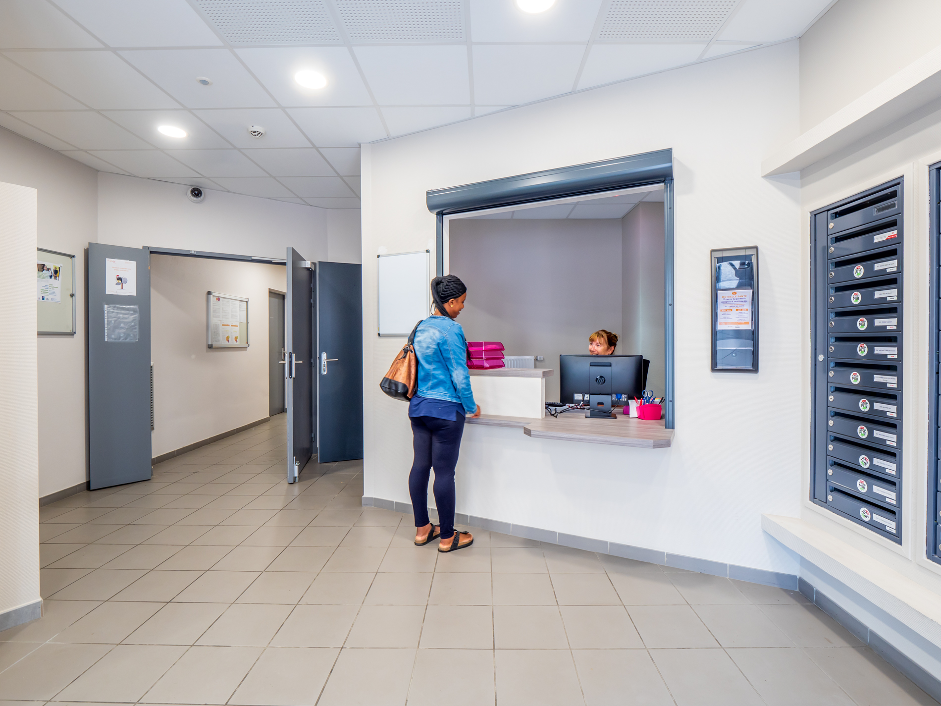 Une jeune fille est dans un sas d&#039;entrée, une personne l&#039;accueille. sur la droite on voit des boites aux lettre et au fond un couloir menant aux chambres.