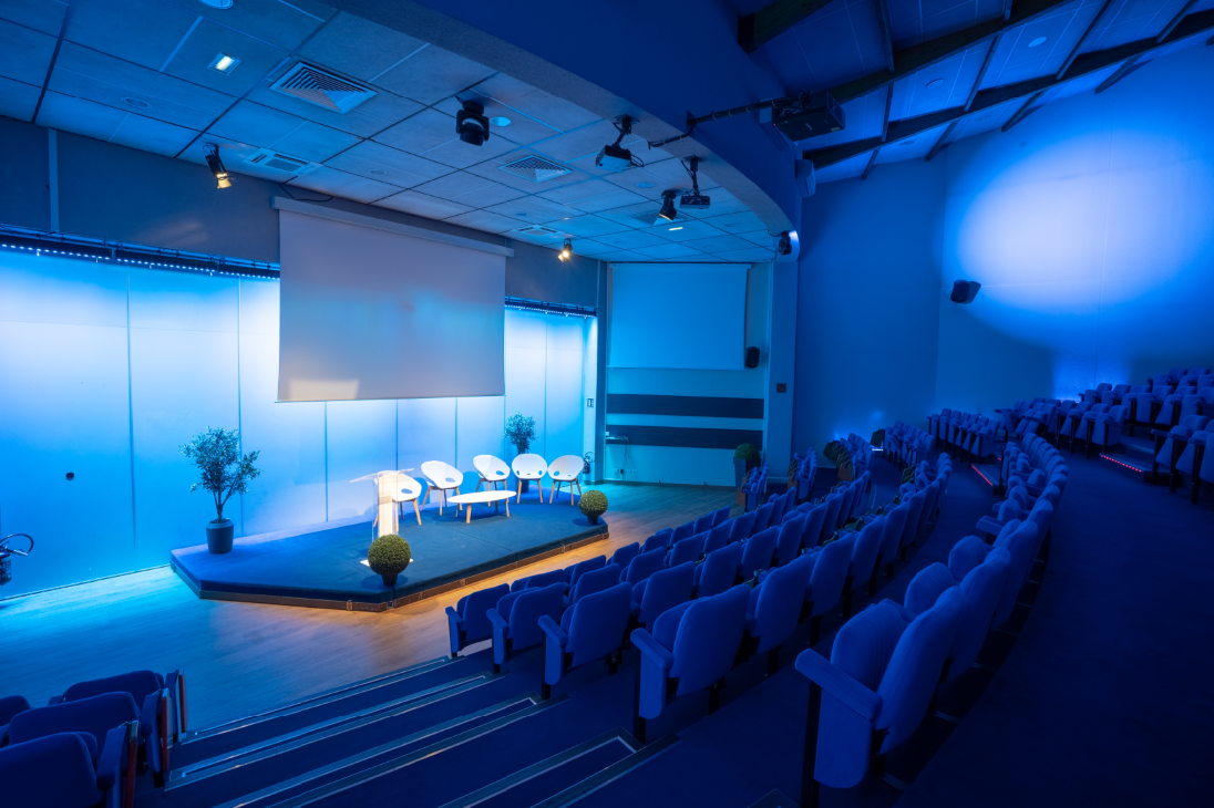 Vue de l’amphithéâtre depuis le haut gauche des gradins avec vue sur la scène éclairée sur laquelle sont disposée 5 fauteuils blancs autour d’une table basse et surmonté d’un écran de vidéo-projection