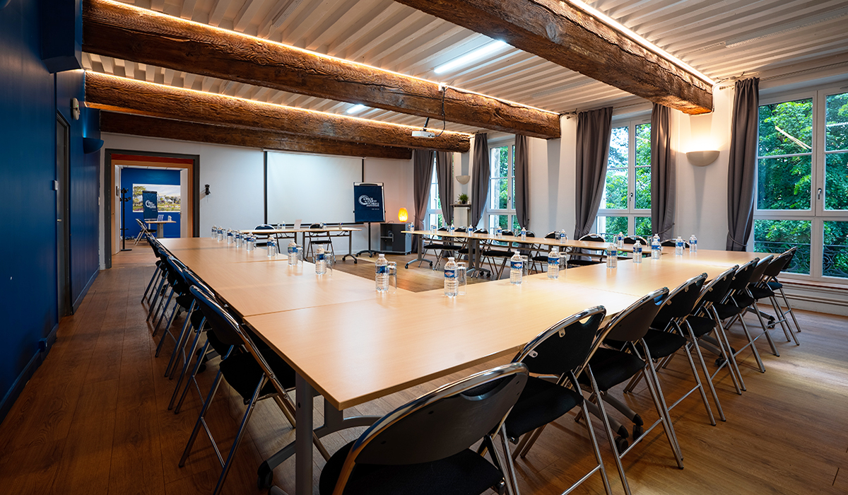 Vue d’une salle de sous-commissions prise du fond de la salle. Les tables sont disposées en U pour une réunion de travail. Les chaises sont disposées tout autour du U et l’on aperçoit au fond l’entrée et le bureau et sur le fond à droite un paperboard.