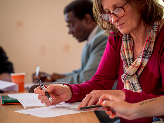 Une dame est en train de remplir un formulaire papier et se fait accompagner par une autre qui lui montre où et comment le renseigner.