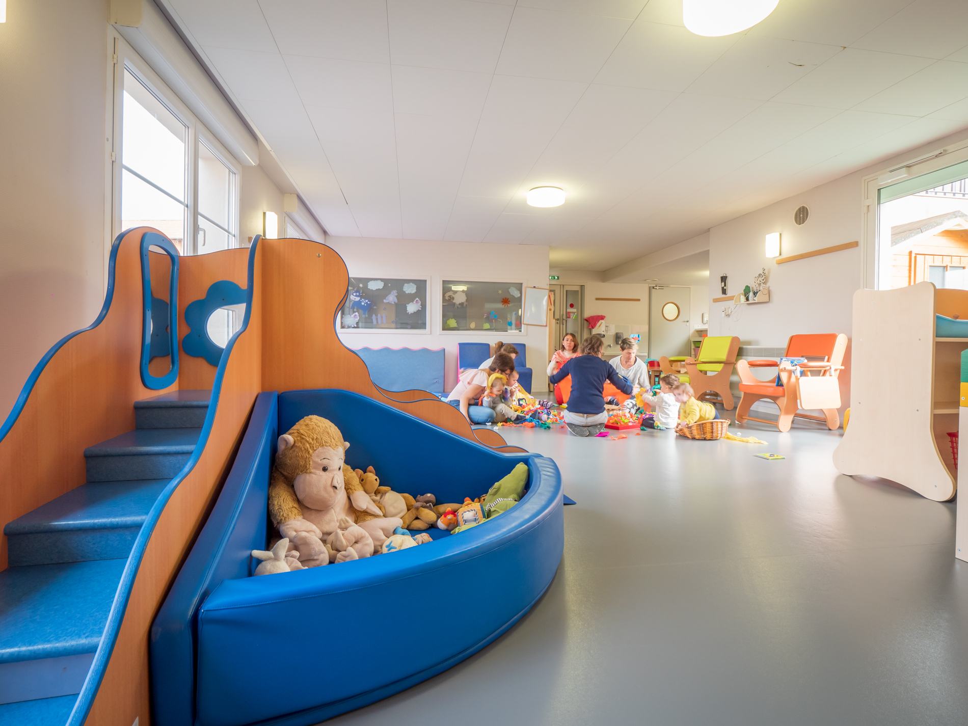 une salle de jeux colorée et lumineuse, on voit en fond un groupe d&#039;adultes et de petits enfants regroupés autour d&#039;une activité