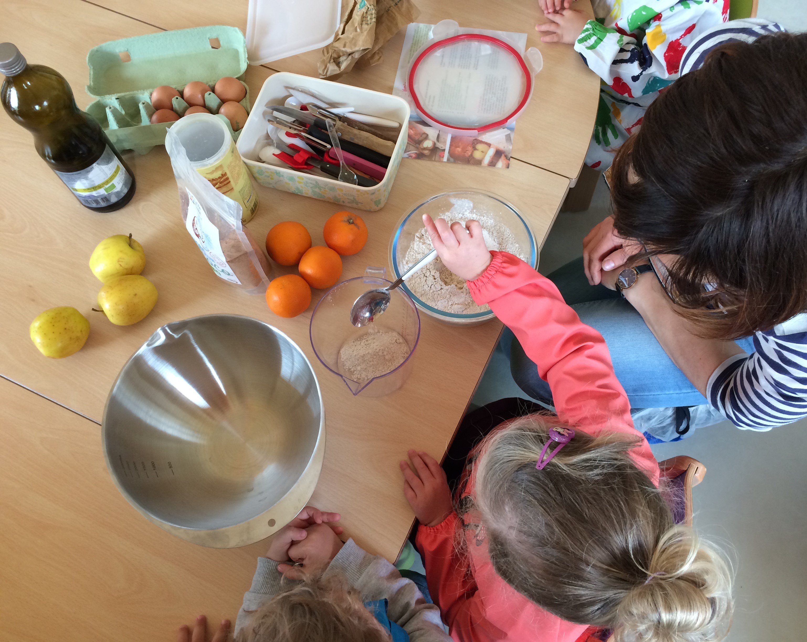 On voit une adulte expliquer à une enfant comment préparer les ingrédient d&#039;un dessert. Oeuf, farine, pommes, saladier sont disposés sur la table