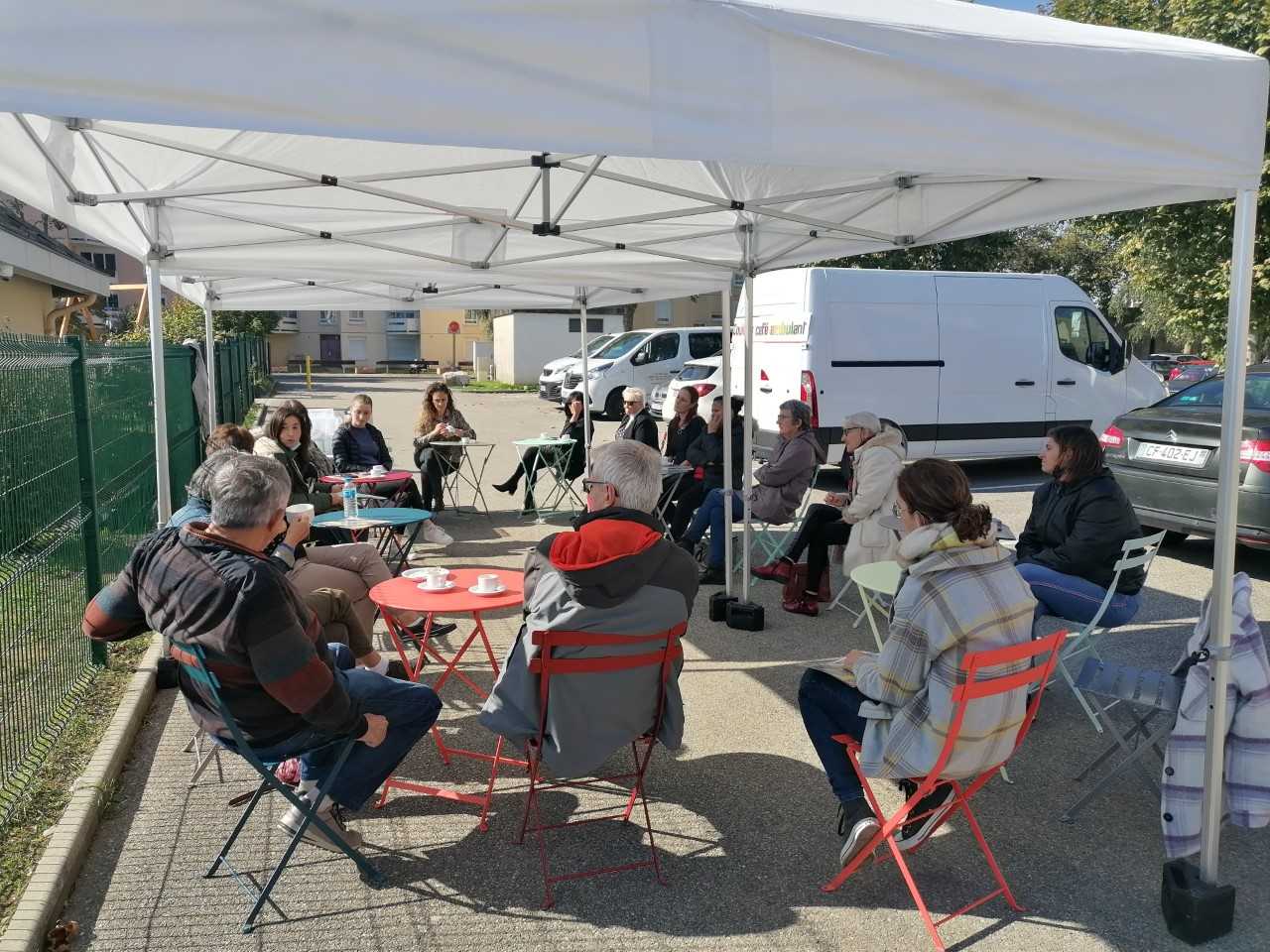 Sous une tente, assis au soleil, des habitants et salariés du centre social se rencontrent et discutent autour d&#039;un café