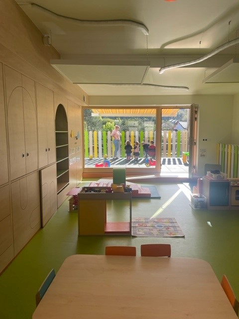 dans une crèche, vue sur l&#039;espace de jeux et une grande baie vitrée donnant sur une terrasse extérieure ensoleillée, où une animatrice et des enfants profitent de l&#039;extérieur