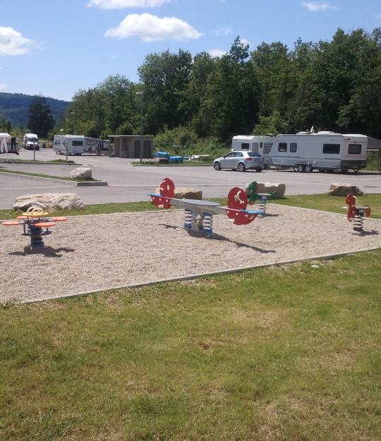 Aire de passage pour les gens du voyage, on voit des jeux aménagés à l'extérieur et des caravanes garées sur un grand parking.