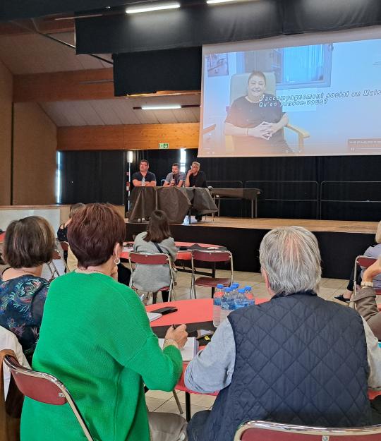 Au premier plan, groups de participants assis à leur table ronde de dos, qui écoutent attentivement les membres d'une autre table ronde sur scène.