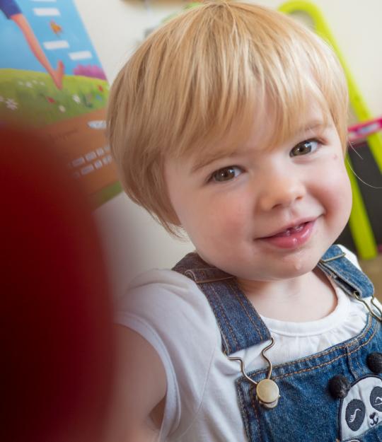 Un jeune enfant blond en salopette bleu regarde l'appareil qui le prend en photo et le touche du doigt avec un sourire