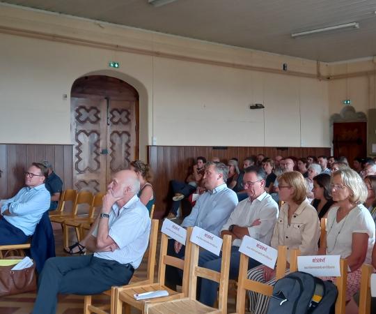 Vue de face : dans le château, les invités assistent à l'assemblée générale, assis sur des chaises en bois disposées en lignes