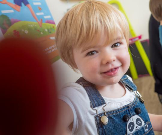 Un jeune enfant blond en salopette bleu regarde l'appareil qui le prend en photo et le touche du doigt avec un sourire