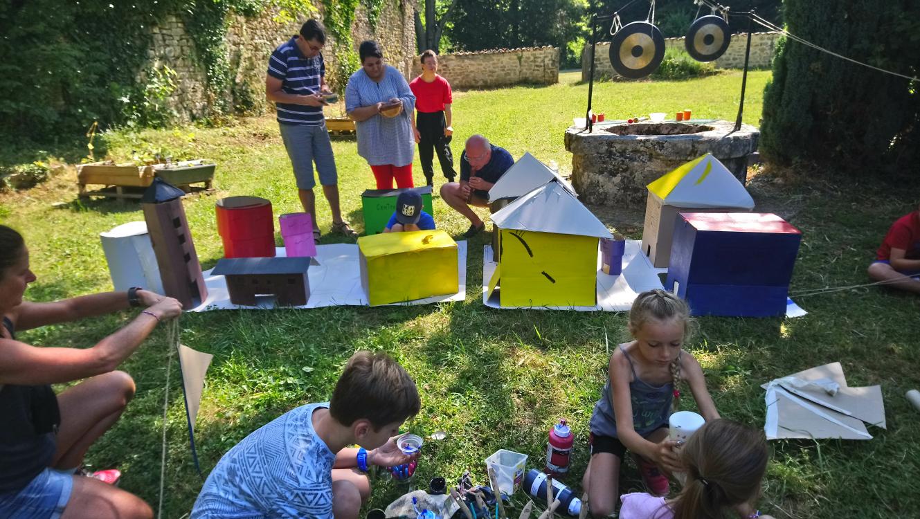 Des enfants et adultes fabriquent des grands objets (maisons, cylindres, …) en carton dans un parc ensoleillé