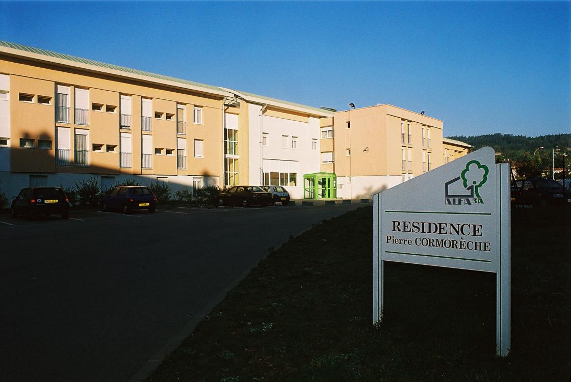 vue du batiment extérieur avec le panneau et la résidence en arrière plan, et des places de parking le long du batiment.