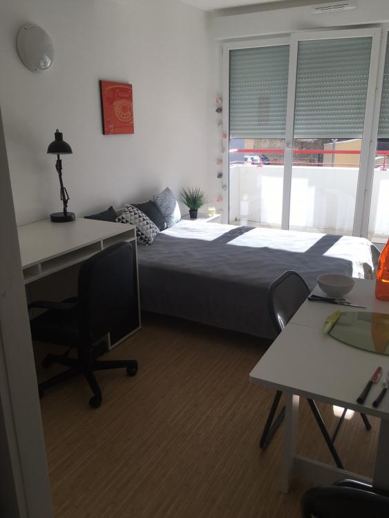 Un studio étudiant : on voit une chambre meublée (lit double, placard, bureau, table, porte-fenêtre) avec son balcon