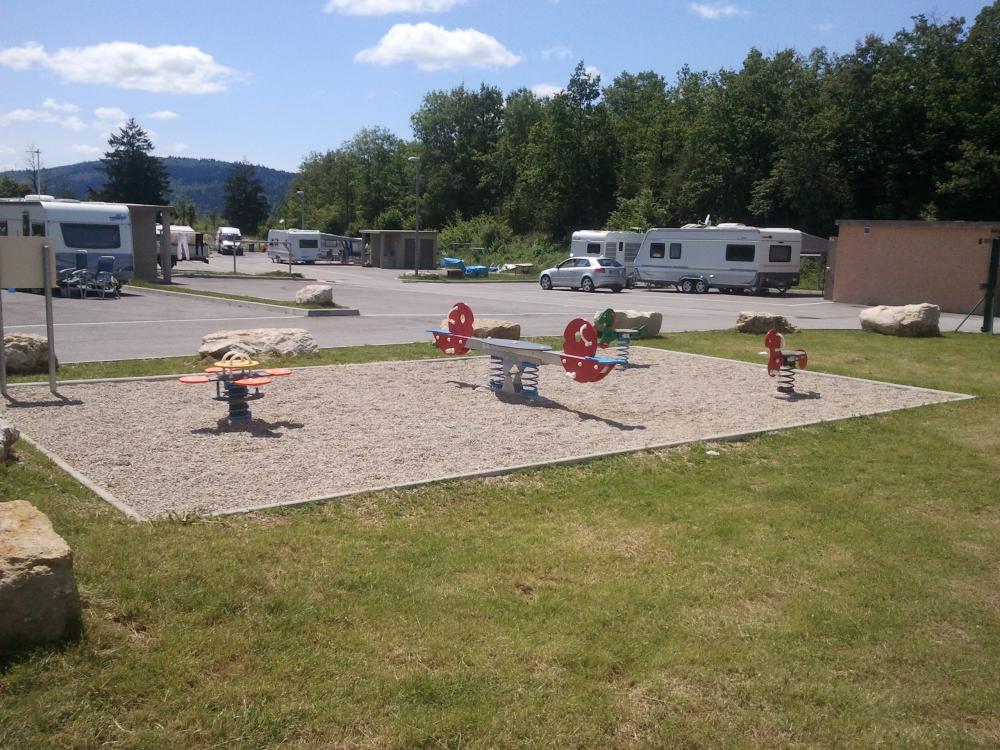 Aire de passage pour les gens du voyage, on voit des jeux aménagés à l'extérieur et des caravanes garées sur un grand parking.