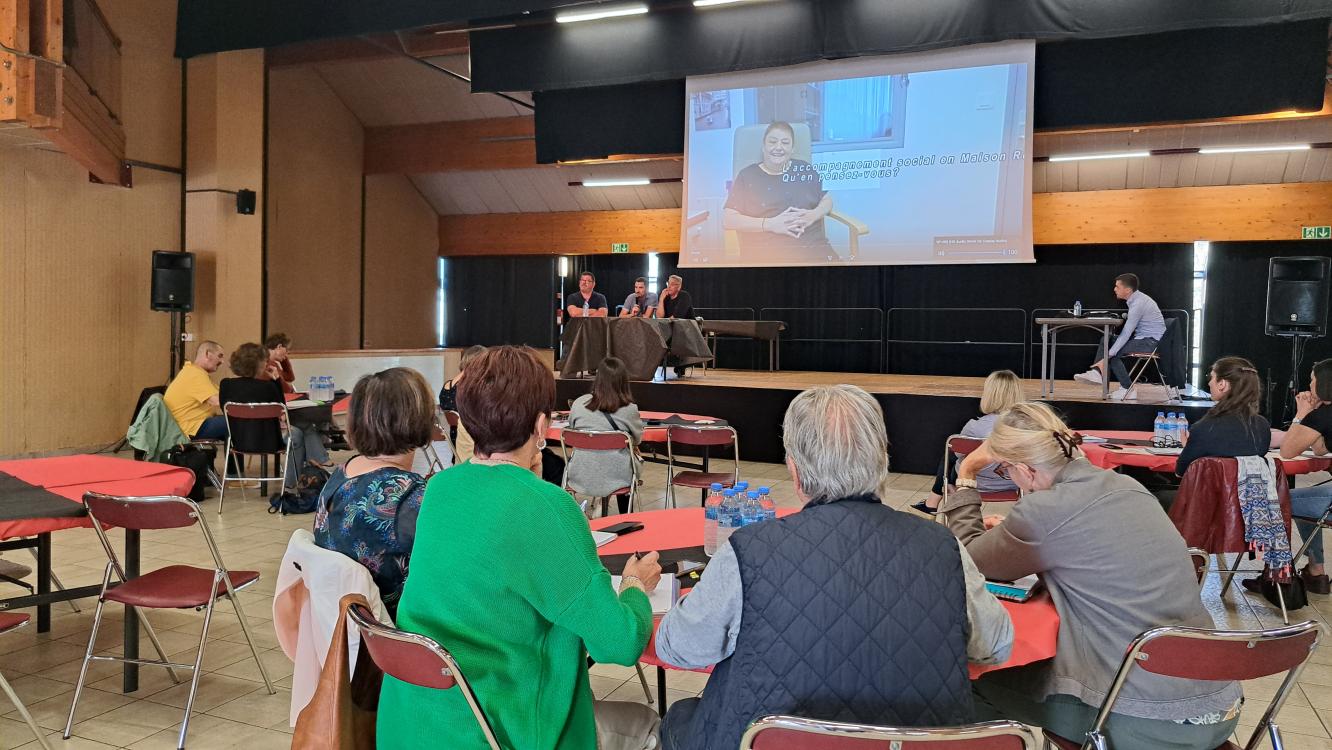 Au premier plan, groups de participants assis à leur table ronde de dos, qui écoutent attentivement les membres d'une autre table ronde sur scène.