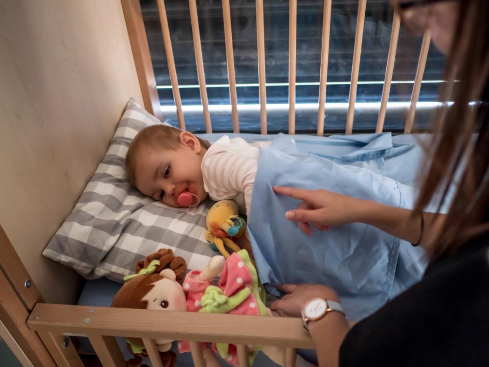 enfant dans son lit à barreau, avec une tétine, et une professionnelle qui pose la main sur lui